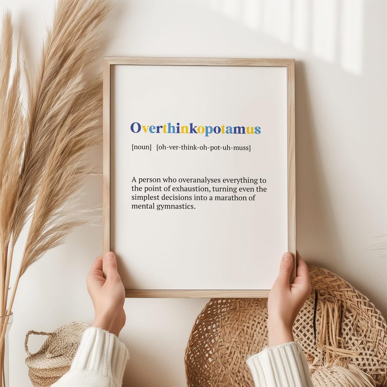 May include: A framed print with a light brown wood frame and a white background. The print features the word "Overthinkopotamus" in blue and yellow letters with a definition below. The definition reads: "[noun] [oh-ver-think-oh-pot-uh-muss] A person who overanalyses everything to the point of exhaustion, turning even the simplest decisions into a marathon of mental gymnastics."