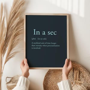 May include: A framed print with the text "In a sec" in a light teal colour. The definition below reads: "A mythical unit of time longer than eternity when procrastination is involved."