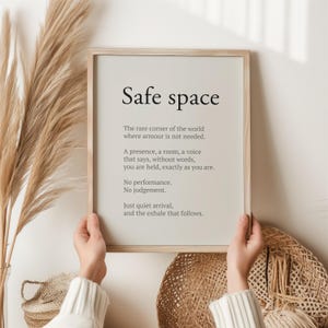 May include: A framed print with the words "Safe space" in a minimalist design. The text includes a description of a safe space, with a neutral background and a light wooden frame. The print is held by a person.