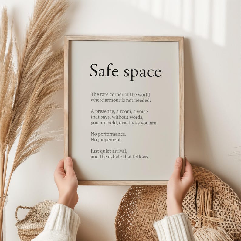 May include: A framed print with the words "Safe space" in a minimalist design. The text includes a description of a safe space, with a neutral background and a light wooden frame. The print is held by a person.