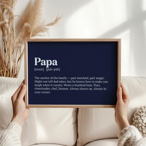 Op de afbeelding: Een ingelijste print met de definitie van "Papa" in witte tekst op een marineblauwe achtergrond. De definitie beschrijft een vaderfiguur als een bron van ondeugd, magie en steun. De lijst is lichtbruin.