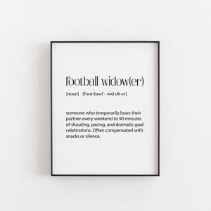 以下が含まれることがあります： ミニマリストフォントで「football widow(er)」というテキストが書かれた黒いフレームプリント。定義は、サッカーの試合中に一時的にパートナーを失う人を説明しています。プリントは白黒です。
