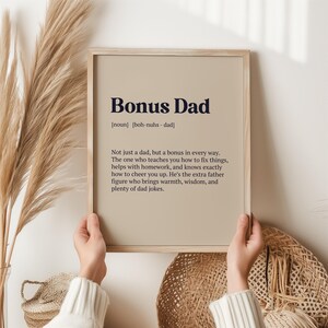 May include: A framed print with a beige background and black text that reads "Bonus Dad [noun] [boh-nuhs - dad] Not just a dad, but a bonus in every way. The one who teaches you how to fix things, helps with homework, and knows exactly how to cheer you up. He's the extra father figure who brings warmth, wisdom, and plenty of dad jokes."