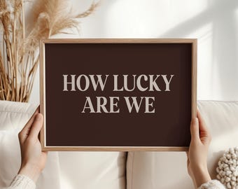 May include: A framed print with the text "HOW LUCKY ARE WE" in a light color against a dark brown background. The frame is a light wood color. The print is held by a person.