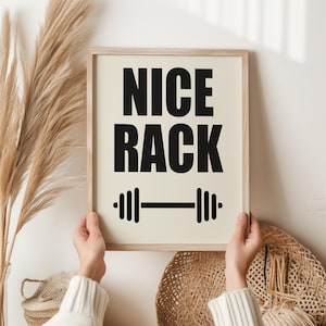 May include: A framed print featuring the text "NICE RACK" in bold black font, with a barbell graphic beneath. The background is cream-colored, and the frame is light wood. The print is held by a person.