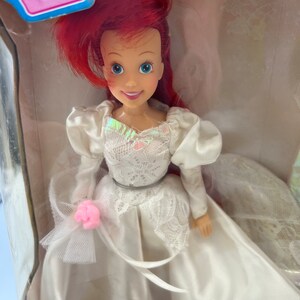 May include: A red-haired doll wearing a white wedding dress with lace detailing and a pink flower. The doll is holding a bouquet of flowers and has a crown on her head.