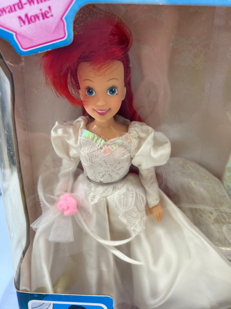 May include: A red-haired doll wearing a white wedding dress with lace detailing and a pink flower. The doll is holding a bouquet of flowers and has a crown on her head.