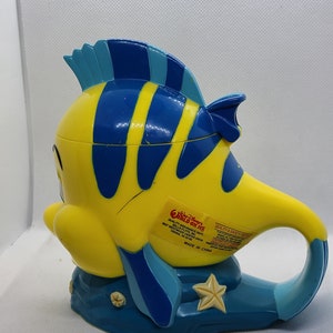 Puede incluir: Una taza de pl&aacute;stico amarilla y azul con forma del personaje Nemo de la pel&iacute;cula Buscando a Nemo. La taza tiene un asa y una base azul con un dise&ntilde;o de estrella de mar. La taza tiene una etiqueta que dice "World on Ice" y "Quality Assurance Dept. Best Westmod Center Drive, Selma, CA 93662. Made in China."