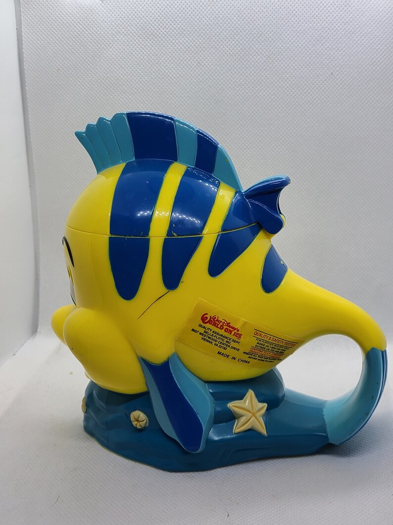 Puede incluir: Una taza de pl&aacute;stico amarilla y azul con forma del personaje Nemo de la pel&iacute;cula Buscando a Nemo. La taza tiene un asa y una base azul con un dise&ntilde;o de estrella de mar. La taza tiene una etiqueta que dice "World on Ice" y "Quality Assurance Dept. Best Westmod Center Drive, Selma, CA 93662. Made in China."