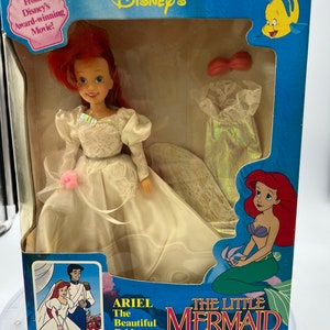 May include: Ariel, the Little Mermaid, is dressed in a white wedding gown with a pink flower. She is holding a bouquet of flowers. The box is blue and features the Disney logo and the words "The Little Mermaid".