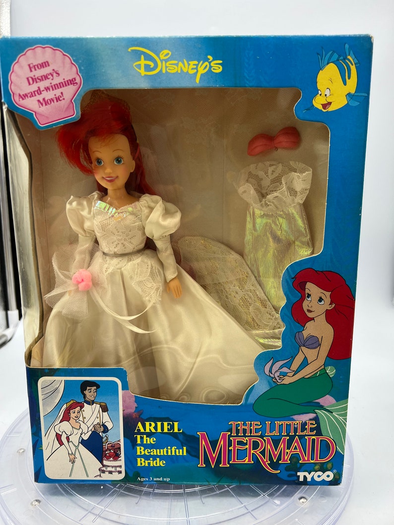 May include: Ariel, the Little Mermaid, is dressed in a white wedding gown with a pink flower. She is holding a bouquet of flowers. The box is blue and features the Disney logo and the words "The Little Mermaid".