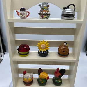 Może przedstawiać: Biała drewniana półka z trzema półkami, na których wystawionych jest dziewięć miniaturowych figurek ceramicznych. Figurki są tematycznie związane z ogrodem i obejmują czajnik, konewkę, czajnik, biedronkę, słońce, ul i trzy kury.