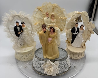 Könnte beinhalten: Eine Sammlung von Vintage-Hochzeitstortenaufsätzen. Die Aufsätze zeigen Miniaturfiguren von Braut und Bräutigam in verschiedenen Posen, umgeben von Spitze, Schleifen und dekorativen Elementen. Die Farbpalette umfasst Elfenbein, Gold und Schwarz.