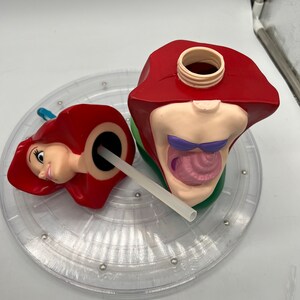 Op de afbeelding: Een rode en groene plastic beker in de vorm van Ariel uit De Kleine Zeemeermin. De beker is open en heeft een rietje dat eruit steekt. De beker staat op een doorzichtige plastic draaitafel.