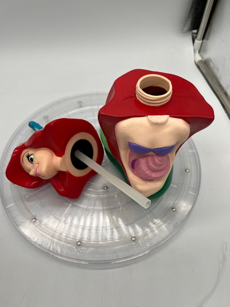 Op de afbeelding: Een rode en groene plastic beker in de vorm van Ariel uit De Kleine Zeemeermin. De beker is open en heeft een rietje dat eruit steekt. De beker staat op een doorzichtige plastic draaitafel.
