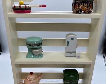 May include: A white wooden shelf unit with three shelves, each displaying a different miniature kitchen appliance. The top shelf has a red and white miniature kitchen appliance with a yellow ball on top. The middle shelf has a green stand mixer and a white refrigerator with a miniature milk carton. The bottom shelf has a pink teapot with a heart on the lid and a green refrigerator with a miniature milk carton.