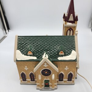 K&ouml;nnte beinhalten: Ein Keramik-Kirchengeb&auml;ude mit einem gr&uuml;nen Dach, einem braunen Turm und Buntglasfenstern. Die Kirche ist mit Schnee verziert und hat einen Kranz an der T&uuml;r.