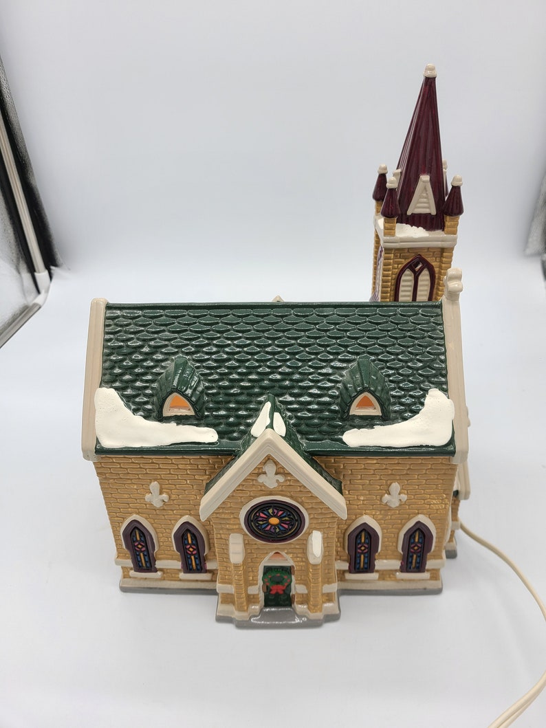 K&ouml;nnte beinhalten: Ein Keramik-Kirchengeb&auml;ude mit einem gr&uuml;nen Dach, einem braunen Turm und Buntglasfenstern. Die Kirche ist mit Schnee verziert und hat einen Kranz an der T&uuml;r.