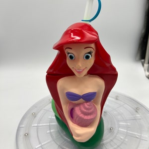 Op de afbeelding: Een rode, groene en paarse plastic beker in de vorm van Ariel, de Kleine Zeemeermin, die een roze schelp vasthoudt. De beker heeft een blauwe en witte rietje.