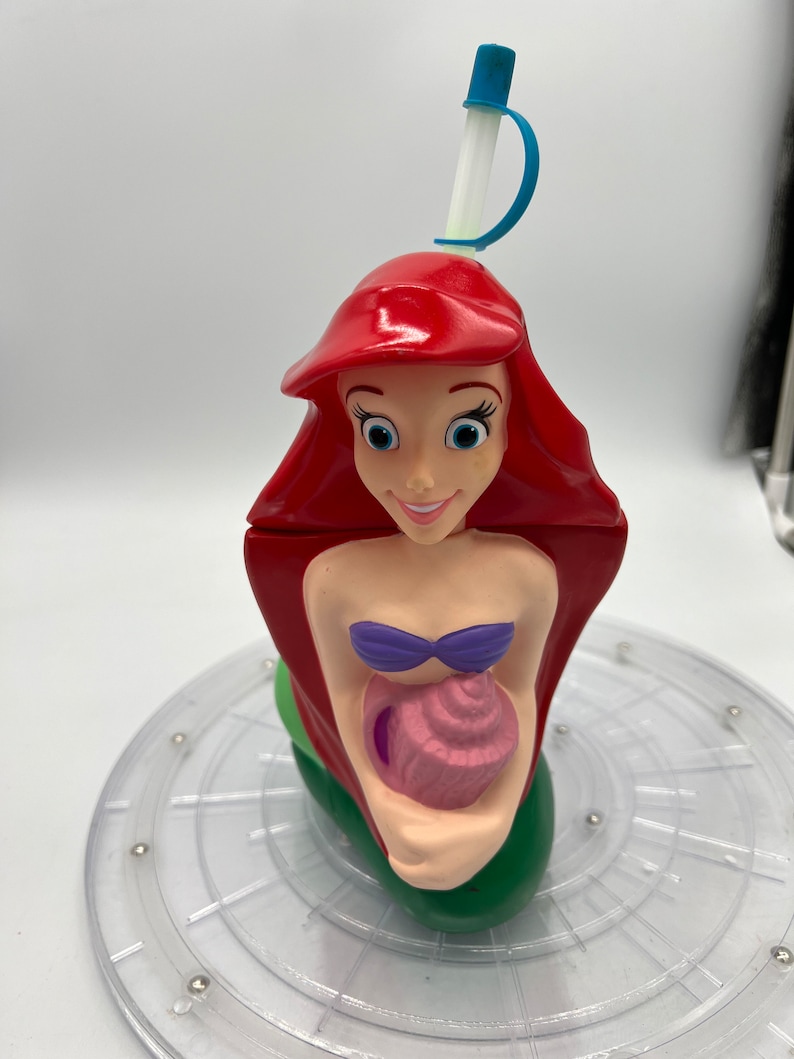 Op de afbeelding: Een rode, groene en paarse plastic beker in de vorm van Ariel, de Kleine Zeemeermin, die een roze schelp vasthoudt. De beker heeft een blauwe en witte rietje.