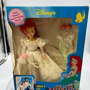May include: Ariel, the Little Mermaid, is dressed in a white wedding gown with a pink flower. She is holding a bouquet of flowers. The box is blue and features the Disney logo and the words "The Little Mermaid".