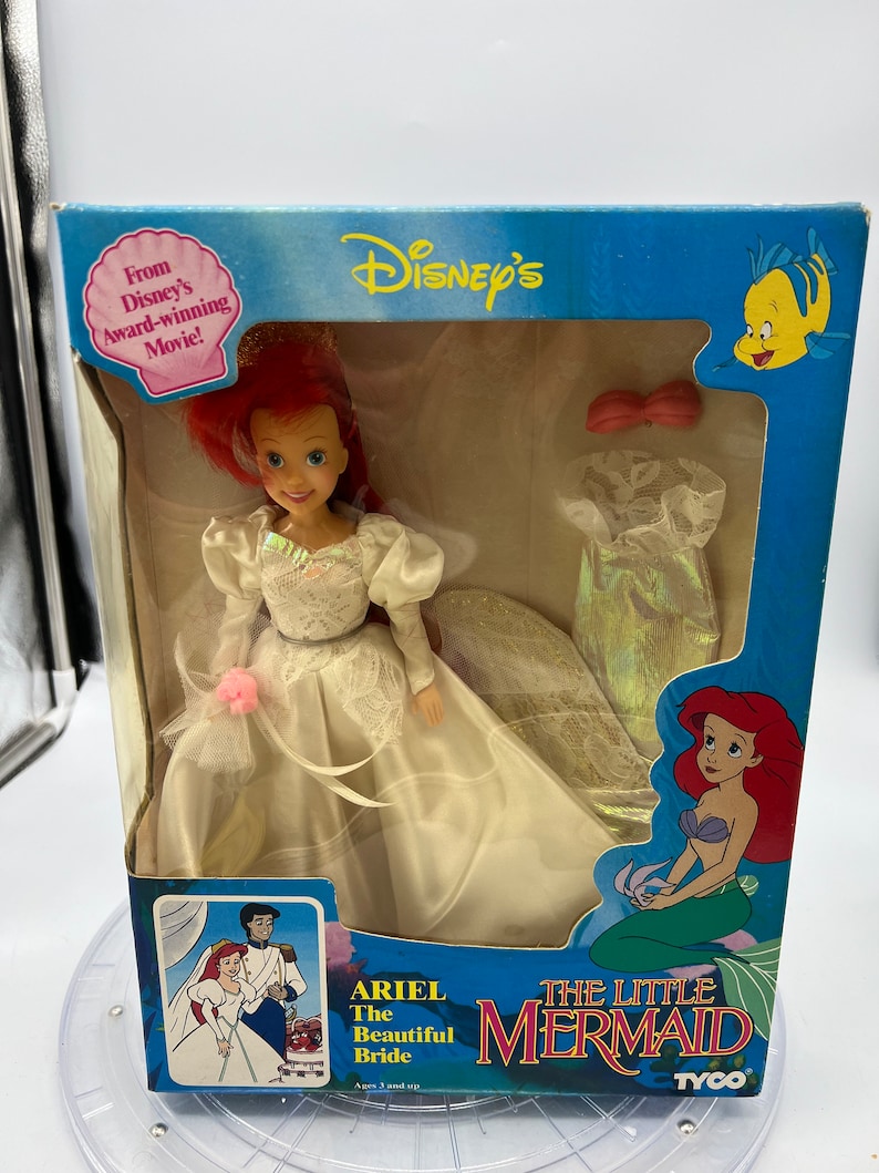 May include: Ariel, the Little Mermaid, is dressed in a white wedding gown with a pink flower. She is holding a bouquet of flowers. The box is blue and features the Disney logo and the words "The Little Mermaid".