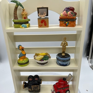 May include: A white wooden shelf unit with three shelves, each displaying a collection of small, colorful ceramic trinket boxes. The boxes feature various themes, including a tropical island, a candy cane, a fruit basket, a duck, a bowl of fruit, a Hollywood award, a movie camera, and a lawnmower.