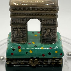 Op de afbeelding: Een miniatuur keramische Arc de Triomphe sculptuur. De boog is een gedempte grijze kleur, met een groene basis versierd met kleurrijke stippen. De basis is een scharnierende doos met een hartvormige sluiting.