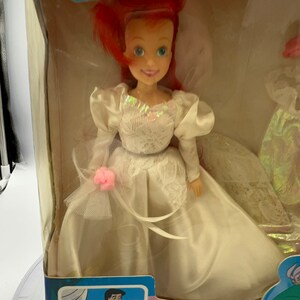 May include: A plastic doll of Ariel from The Little Mermaid wearing a white wedding dress with a pink flower on the side. The doll is in a box with a picture of Ariel and Prince Eric on the front. The box also says "Ariel The Beautiful Bride" and "The Little Mermaid".
