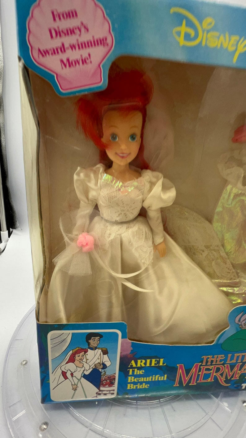 May include: A plastic doll of Ariel from The Little Mermaid wearing a white wedding dress with a pink flower on the side. The doll is in a box with a picture of Ariel and Prince Eric on the front. The box also says "Ariel The Beautiful Bride" and "The Little Mermaid".