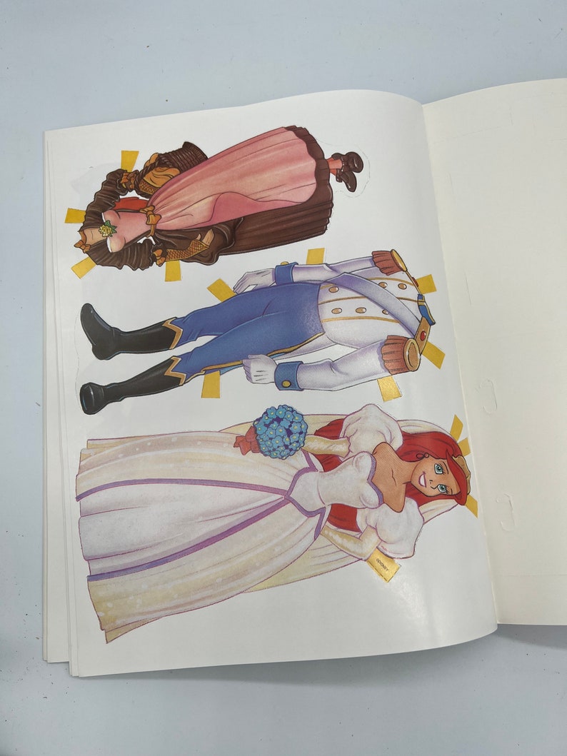 Puede incluir: Conjunto de mu&ntilde;ecas de papel con una novia con un vestido blanco y un ramo de flores azules, un pr&iacute;ncipe con un uniforme azul y blanco y una mujer con un vestido rosa y marr&oacute;n.