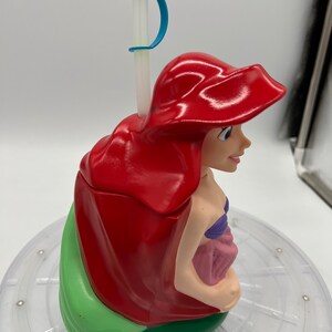Op de afbeelding: Een rode en groene plastic beker in de vorm van Ariel uit De Kleine Zeemeermin. De beker heeft een rietje met een blauwe bovenkant en een blauwe handgreep.