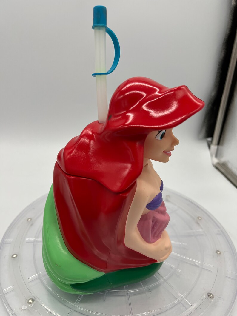 Op de afbeelding: Een rode en groene plastic beker in de vorm van Ariel uit De Kleine Zeemeermin. De beker heeft een rietje met een blauwe bovenkant en een blauwe handgreep.