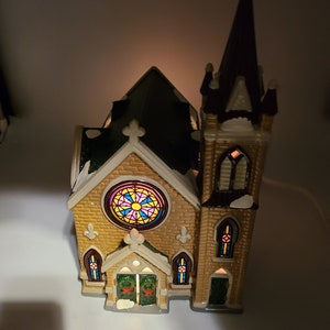 K&ouml;nnte beinhalten: Eine kleine Keramikkirche mit braunem Dach, einem Buntglasfenster und einem beleuchteten Innenraum. Die Kirche hat einen hohen Turm mit braunem Dach und einer wei&szlig;en, schneebedeckten Spitze.