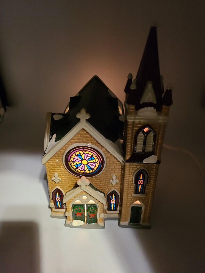 K&ouml;nnte beinhalten: Eine kleine Keramikkirche mit braunem Dach, einem Buntglasfenster und einem beleuchteten Innenraum. Die Kirche hat einen hohen Turm mit braunem Dach und einer wei&szlig;en, schneebedeckten Spitze.