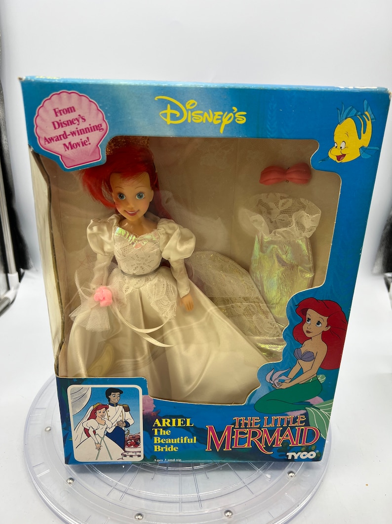 May include: A plastic Ariel doll from Disney's The Little Mermaid wearing a white wedding dress with a pink flower. The doll is packaged in a box with a picture of Ariel and Prince Eric on the front. The box also features the text "Ariel The Beautiful Bride" and "The Little Mermaid".