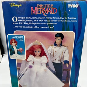 May include: A box for a Disney's The Little Mermaid wedding doll set. The box features Ariel and Eric in their wedding attire. The text on the box reads "Disney's The Little Mermaid" and "Once upon a time, in the kingdom beneath the sea, lived the beautiful mermaid princess, Ariel. Then one day she met the handsome human prince, Eric! They fell deeply in love and got married...and what a beautiful wedding ceremony it was!"