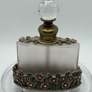 May include: A frosted glass perfume bottle with a crystal stopper and a floral design around the base. The bottle is sitting on a clear plastic base.