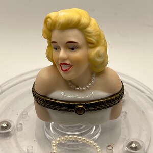 May include: A porcelain figurine of a woman with blonde hair wearing a white dress and a pearl necklace. The figurine is on a clear plastic base with a pearl necklace.