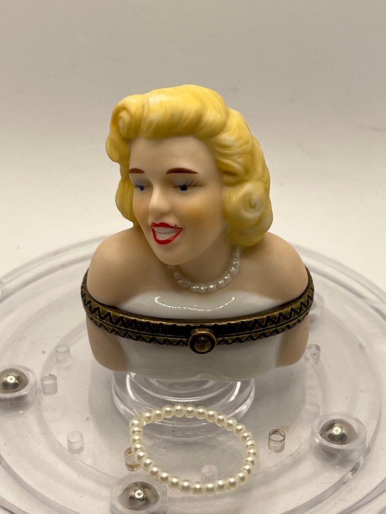 May include: A porcelain figurine of a woman with blonde hair wearing a white dress and a pearl necklace. The figurine is on a clear plastic base with a pearl necklace.