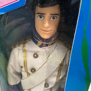 Pode incluir: Uma boneca de pl&aacute;stico do Pr&iacute;ncipe Eric do filme da Disney A Pequena Sereia. Ele est&aacute; vestindo um uniforme branco com detalhes dourados e uma faixa branca. Ele tem cabelo castanho e olhos azuis.