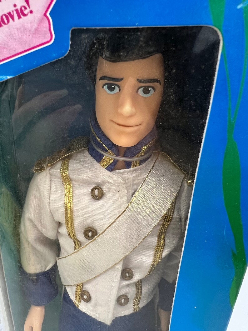 Pode incluir: Uma boneca de pl&aacute;stico do Pr&iacute;ncipe Eric do filme da Disney A Pequena Sereia. Ele est&aacute; vestindo um uniforme branco com detalhes dourados e uma faixa branca. Ele tem cabelo castanho e olhos azuis.