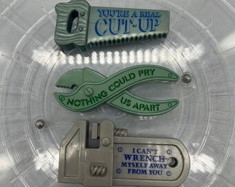 Op de afbeelding: Drie grijze gereedschapvormige magneten met blauwe en groene tekst. De bovenste magneet is een zaag met de tekst "YOU'RE A REAL CUT-UP". De middelste magneet is een tang met "NOTHING COULD PRY US APART". De onderste magneet is een moersleutel met "I CAN'T WRENCH MYSELF AWAY FROM YOU."