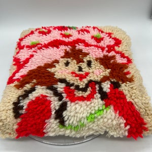 Op de afbeelding: Een vintage naaldviltkussen met een cartoon aardbei-personage met bruin haar, een roze hoed en een rode jurk. Het kussen is gemaakt van wit, roze, rood, bruin en groen garen.