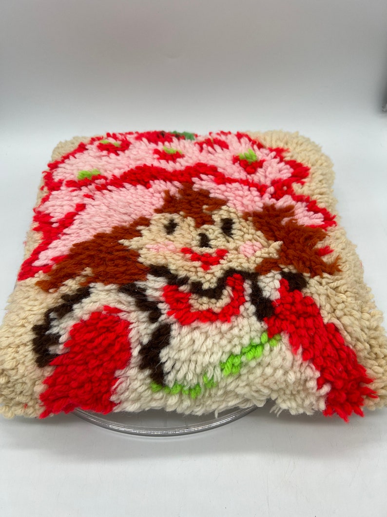 Op de afbeelding: Een vintage naaldviltkussen met een cartoon aardbei-personage met bruin haar, een roze hoed en een rode jurk. Het kussen is gemaakt van wit, roze, rood, bruin en groen garen.