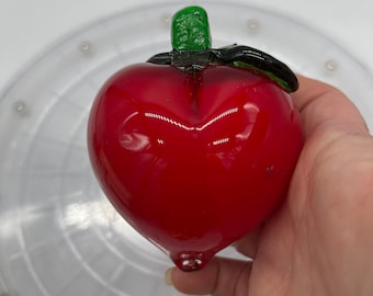 May include: A red glass apple with a green stem and a black leaf. The apple is held in a hand and has a glossy finish. The apple is a decorative item.