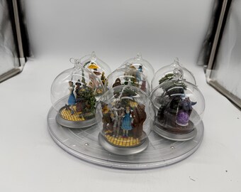 Peut inclure: Un ensemble de six boules de Noël en verre transparent, chacune contenant une scène miniature avec des figurines du Magicien d'Oz. Les scènes représentent des personnages comme Dorothy, l'Homme de Fer, l'Épouvantail et le Lion sur une route de briques jaunes.