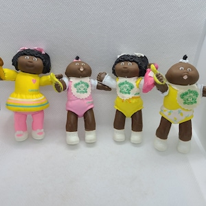 Op de afbeelding: Vier Cabbage Patch Kids poppen met bruine huid, zwart haar en verschillende outfits. De poppen dragen gele, roze en witte kleding met groene accenten. De poppen houden verschillende accessoires vast, waaronder een lepel, een fles en een kam.