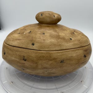 May include: A tan ceramic lidded container shaped like a potato with a textured surface, featuring dark brown speckles and an oval form. The lid includes a small, rounded handle at the top, mimicking the appearance of a natural potato skin.