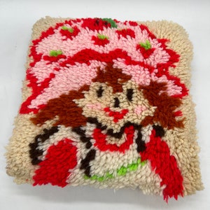 Op de afbeelding: Een vintage naaldviltkussen met een cartoonfiguur met rood haar, een roze hoed en een rood hart. Het figuur is omringd door wit, rood en groen garen.